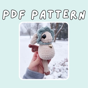 May include: A crocheted bunny doll with a blue bonnet and a heart-shaped charm. The doll is beige with brown accents and is held against a snowy background. The text "PDF PATTERN" is at the top of the image.