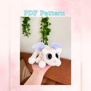 May include: A crocheted white dragon with purple wings and pink accents. The dragon has large, colorful eyes and a pink nose. The dragon is being held in a person's hand. PDF Pattern.