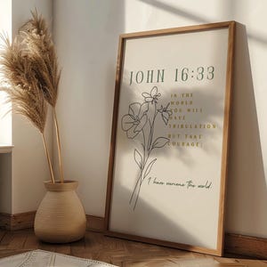 May include: A framed print featuring the text "JOHN 16:33" and a floral illustration. The text reads "In the world you will have tribulation but take courage; I have overcome the world." The frame is wood-toned, and the background is off-white.