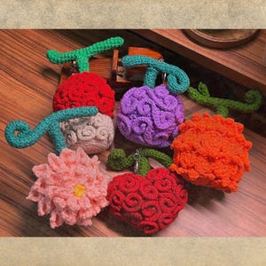 Op de afbeelding: Zes gebreide bloemvormige ornamenten in verschillende kleuren, waaronder rood, roze, oranje, paars en wit. Elk ornament heeft een steel met een groen gebreid blad.