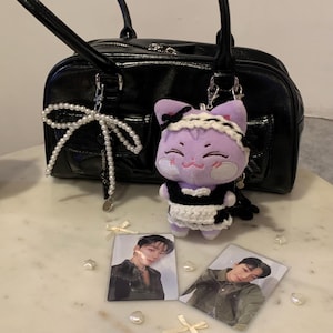 May include: A black patent leather handbag with silver hardware, a pearl bow charm, and a purple cat plushie wearing a maid outfit. Two photo cards of a person are on the table.