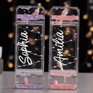 Könnte beinhalten: Zwei transparente, rechteckige Flüssigkeits-Bewegungsspielzeuge mit den Namen "Sophia" und "Amilia" in weißer Schrift. Sie zeigen eine Hasenfigur und schwebenden Glitzer. Eines hat lila und das andere rosa Akzente.