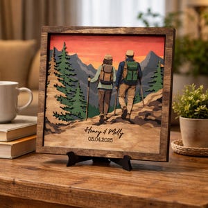 Könnte beinhalten: Holzgerahmtes Kunstwerk, das ein Paar beim Wandern in den Bergen bei Sonnenuntergang zeigt. Das Kunstwerk ist mehrschichtig mit einem braunen Rahmen, und die Namen "Henry & Molly" und das Datum "03.04.2025" sind unten eingraviert. Das Kunstwerk wird auf einem schwarzen Ständer ausgestellt.