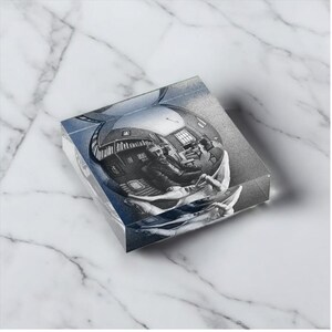 May include: A clear, square-shaped glass paperweight with a black and white illustration of a room with a person inside a sphere. The paperweight has a blue tint and sits on a white marble surface.