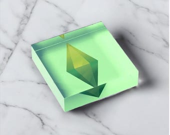 May include: A square, translucent, light green object with a yellow and green diamond-shaped design in the center. The object is made of a solid material and sits on a white and gray marble surface.