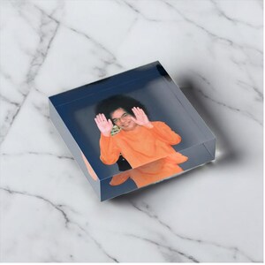 May include: A clear acrylic block featuring a photograph of a person in an orange outfit with their hands up. The background is a dark blue color. The block is sitting on a white and gray marble surface.