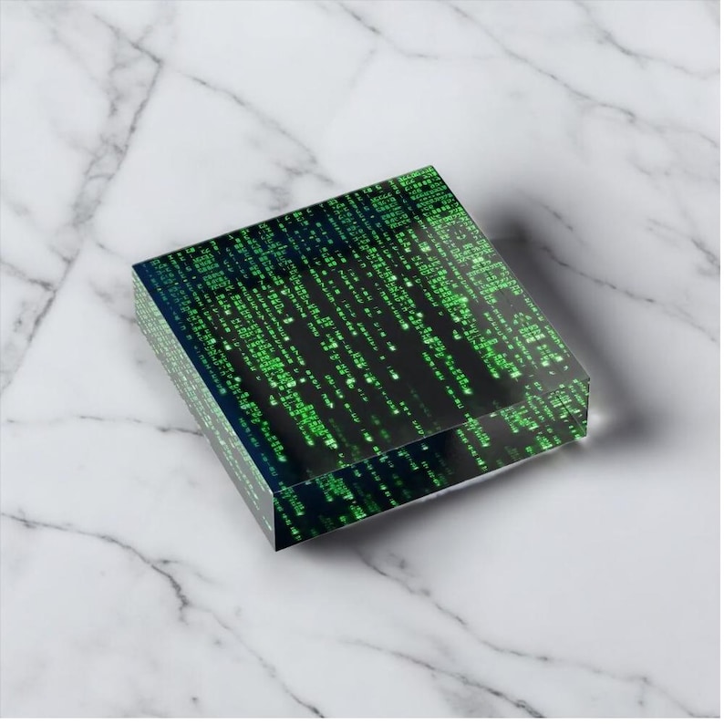 May include: A clear, rectangular block featuring a digital rain pattern in shades of green against a black background. The design resembles binary code or a computer matrix. The block is displayed on a white marble surface.