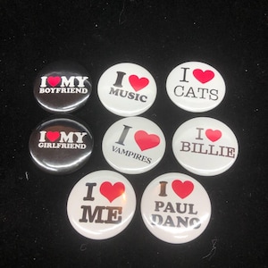 Puede incluir: Ocho pines redondos con corazones rojos y texto negro. Los pines dicen "Amo a mi novio", "Amo a mi novia", "Amo la música", "Amo los vampiros", "Amo los gatos", "Amo a Billie", "Amo a mí mismo", y "Amo a Paul dano".