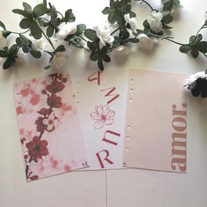 May include: Three rectangular planner inserts with punched holes along the left side. The first features red cherry blossoms, the second has the word AMOR written vertically with a flower, and the third has the word amor printed vertically.