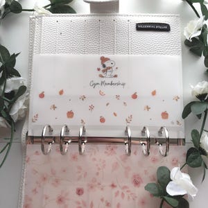 May include: A white textured binder featuring a black label reading MILLENNIAL DOLLAR. Inside, a translucent divider displays a Snoopy graphic, the text Gym Membership, and autumn motifs. Below, a floral patterned sheet with pink roses.