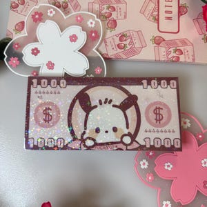 May include: A decorative sticker resembling a $1000 bill featuring a cartoon dog, surrounded by glitter. The sticker is placed on a white surface with a pink floral design and a notebook with strawberry illustrations.