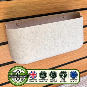 Puede incluir: Un bolsillo de almacenamiento de fieltro gris claro con esquinas redondeadas y dos cierres circulares, montado sobre una superficie de listones de madera horizontales. La imagen incluye un logotipo con el texto Hook In Elements, Designed in the UK, 3D Printed In-House, Tried & Tested, Fits Hymer y Ships to UK & EU.