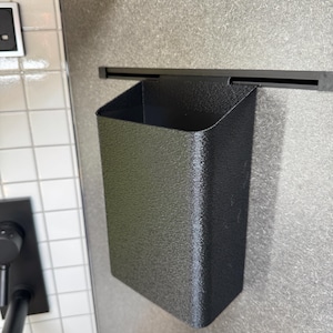 Puede incluir: Un contenedor de almacenamiento rectangular de color negro mate con una superficie texturizada, diseñado para colgarse de un riel horizontal negro. El contenedor cuenta con una parte superior abierta y un gancho en la parte posterior.