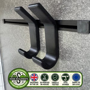 Op de afbeelding: Twee zwarte J-vormige haken gemonteerd op een horizontale zwarte rail. De haken hebben een gebogen ontwerp voor het ophangen van voorwerpen. Een cirkelvormig logo onderaan bevat de tekst Hook In Elements, Designed In The UK, 3D Printed In-House, Tried & Tested, Fits Hymer en Ships To UK & EU.
