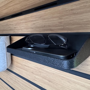 Könnte beinhalten: Ein schwarzes, rechteckiges Wandregal mit strukturierter Oberfläche, auf dem eine schwarze Sonnenbrille und ein schwarzes Smartphone liegen. Das Regal ist an einer hellbraunen Holzpaneelwand mit horizontalen schwarzen Rillen und einer grauen Filztasche befestigt.