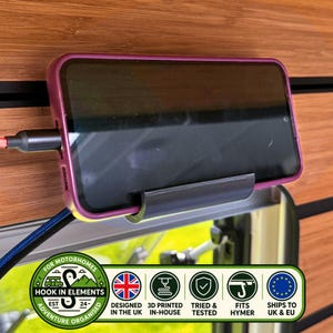 Op de afbeelding: Een zwarte plastic smartphonehouder bevestigd aan een houten wand, met daarin een smartphone met een paarse hoes. Een zwarte oplaadkabel is aangesloten op de zijkant van het toestel. Een rechthoekig label met tekst en iconen is eronder zichtbaar.