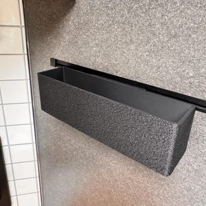 Może przedstawiać: Czarny prostokątny pojemnik do przechowywania z fakturowaną powierzchnią jest zamontowany na czarnej metalowej szynie. Pojemnik wisi na szarej ścianie.