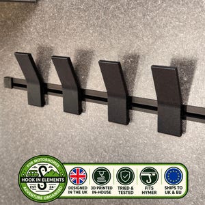 Op de afbeelding: Een zwarte wandrail met vier schuine haken. De rail bevat een logo met de tekst "HOOK IN ELEMENTS EST 24", "DESIGNED IN THE UK", "3D PRINTED IN-HOUSE", "TRIED & TESTED", "FITS HYMER" en "SHIPS TO UK & EU" met bijbehorende iconen.
