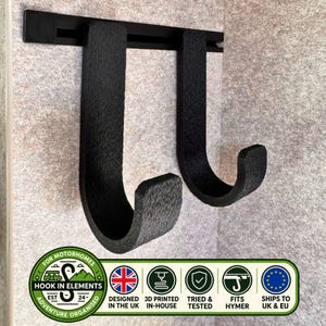 Könnte beinhalten: Ein schwarzes Wandregal mit zwei J-förmigen Haken an einer horizontalen Schiene. Die strukturierten Haken dienen der Organisation. Ein Logo am unteren Rand enthält den Text Hook in Elements, Designed in the UK, 3D Printed In-House, Tried & Tested, Fits Hymer und Ships to UK & EU.
