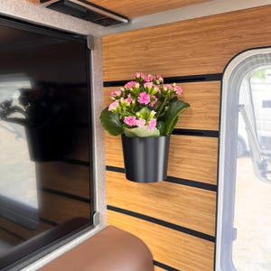 Könnte beinhalten: Eine leuchtend rosa Kalanchoe-Pflanze mit grünen Blättern in einem mattschwarzen zylindrischen Topf, montiert an einer horizontalen Holzpaneelwand mit schwarzen eingelassenen Schienen zwischen den Holzlatten für einen modernen Look.