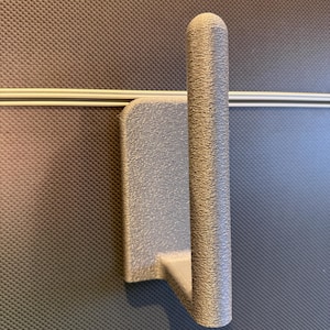 Könnte beinhalten: Ein silberfarbener Wandhaken mit einer texturierten, matten Oberfläche. Der Haken besteht aus einer rechteckigen Montageplatte und einem einzelnen, zylindrischen, nach oben gebogenen Arm mit abgerundeter Spitze zum Aufhängen von Gegenständen.