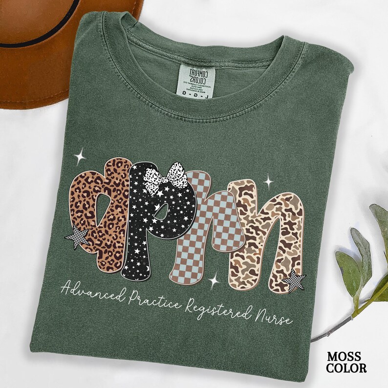 May include: A moss green t-shirt featuring the letters "APRN" in a decorative style. The letters are patterned with leopard print, stars, and a checkered design. The text "Advanced Practice Registered Nurse" is printed below the letters.