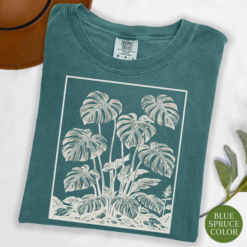 May include: Teal-colored t-shirt featuring a white rectangular graphic of a monstera plant. The plant illustration includes large, detailed leaves and stems. The shirt has a crew neck and short sleeves. The text "BLUE SPRUCE COLOR" is on a green circle.