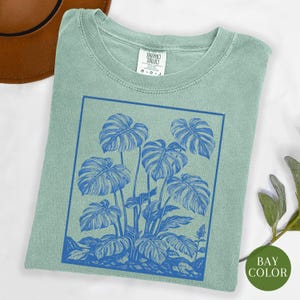 May include: A light green t-shirt featuring a blue illustration of monstera leaves within a blue rectangular frame. The shirt has a crew neck and a tag at the neckline. The words "BAY COLOR" are on a green circle.