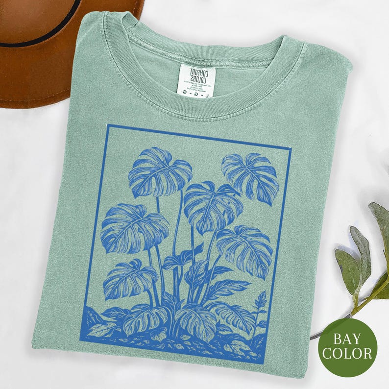 May include: A light green t-shirt featuring a blue illustration of monstera leaves within a blue rectangular frame. The shirt has a crew neck and a tag at the neckline. The words "BAY COLOR" are on a green circle.