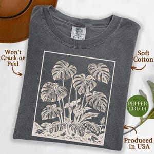 May include: A pepper-colored t-shirt featuring a white framed illustration of monstera leaves. The shirt is made of soft cotton and is produced in the USA. The design won't crack or peel.
