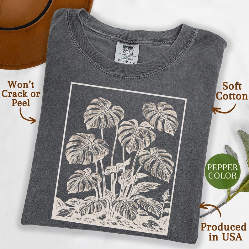 May include: A pepper-colored t-shirt featuring a white framed illustration of monstera leaves. The shirt is made of soft cotton and is produced in the USA. The design won't crack or peel.