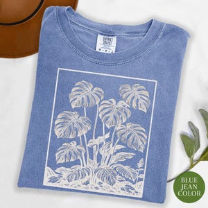 May include: A blue jean color t-shirt with a white rectangular graphic of a monstera plant. The shirt has a crew neck and short sleeves. The tag on the shirt reads "COMFORT COLORS". A green circle with the text "BLUE JEAN COLOR" is in the bottom right corner.