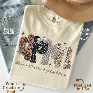 May include: An ivory-coloured t-shirt with the letters "APRN" in a decorative font, featuring leopard, star, and checkered patterns. The text "Advanced Practice Registered Nurse" is printed below. The shirt is made of soft cotton and produced in the USA.