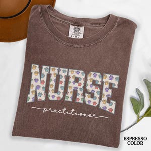 May include: Espresso-colored t-shirt with the word "NURSE" in large, colorful paw print letters. The word "practitioner" is written in cursive below the word "NURSE". The shirt has a crew neck and short sleeves.