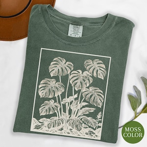 May include: A moss green t-shirt featuring a white rectangular graphic of a monstera plant. The plant illustration includes large, detailed leaves and stems. The shirt has a crew neck and short sleeves.