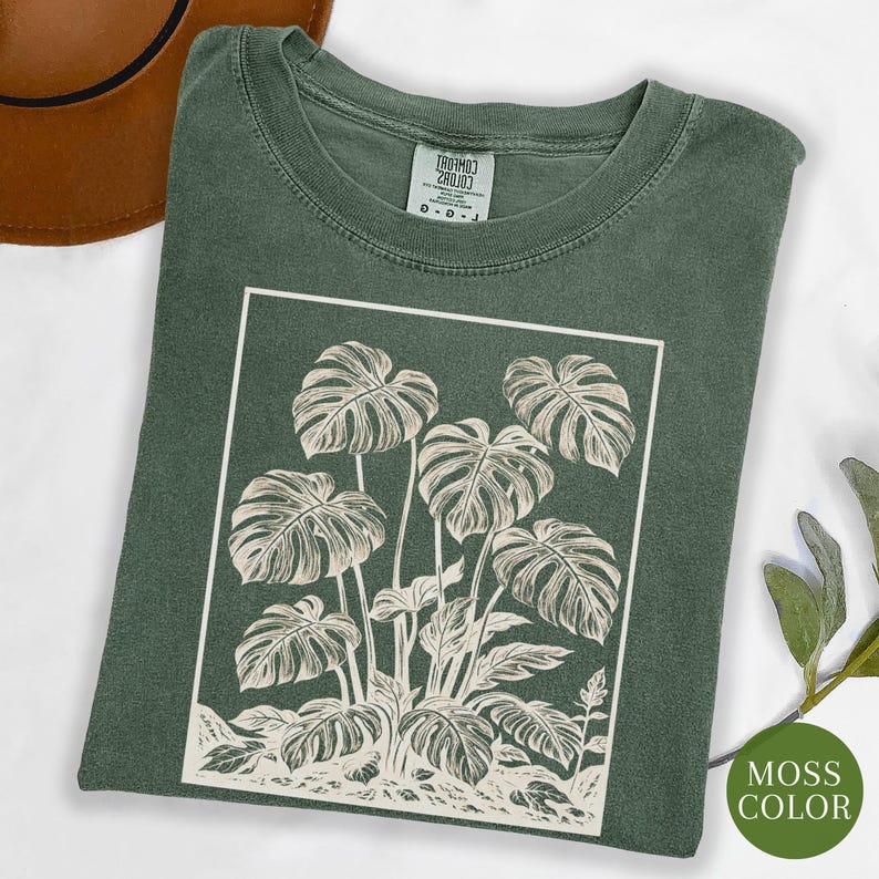 May include: A moss green t-shirt featuring a white rectangular graphic of a monstera plant. The plant illustration includes large, detailed leaves and stems. The shirt has a crew neck and short sleeves.
