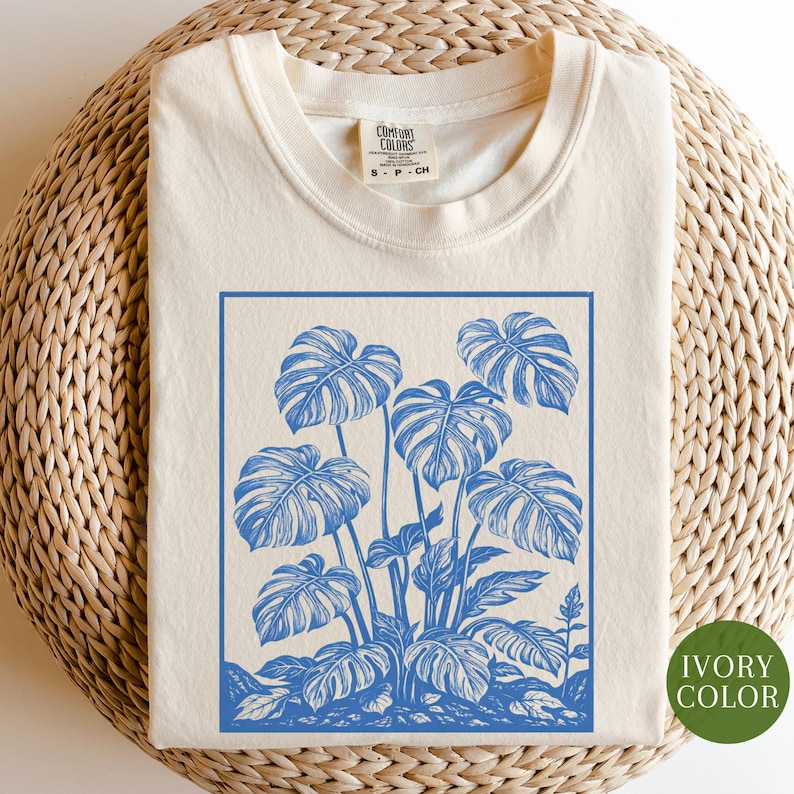 May include: Ivory-colored t-shirt with a blue botanical illustration of monstera leaves. The design is framed within a blue border. The shirt has a crew neck and short sleeves. The tag reads "Comfort Colors".