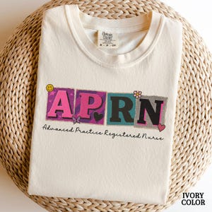 May include: Ivory-colored t-shirt with the letters APRN in a colorful, block-letter design. The shirt includes the text "Advanced Practice Registered Nurse" and decorative hearts, butterflies, and flowers. The shirt is made of a soft material.