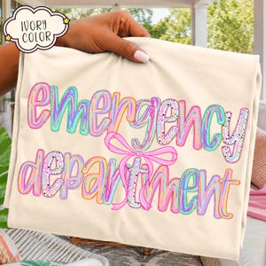 May include: An ivory-colored t-shirt featuring the text "emergency department" in a multi-colored, iridescent, bubble-letter style font. A bright pink ribbon graphic is positioned in the center, tied around the letter "t" in department.