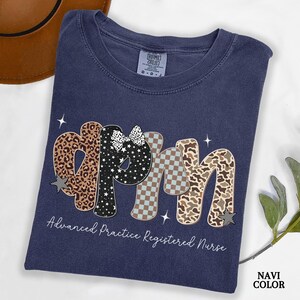 May include: Navy blue t-shirt with the letters "APRN" in a decorative font. The letters feature leopard print, stars, and a checkered pattern. The text "Advanced Practice Registered Nurse" is below the letters.