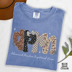 May include: A blue jean coloured t-shirt featuring the letters "APRN" in a decorative style. The letters are patterned with leopard print, stars, and a checkered design. The text "Advanced Practice Registered Nurse" is below the letters.