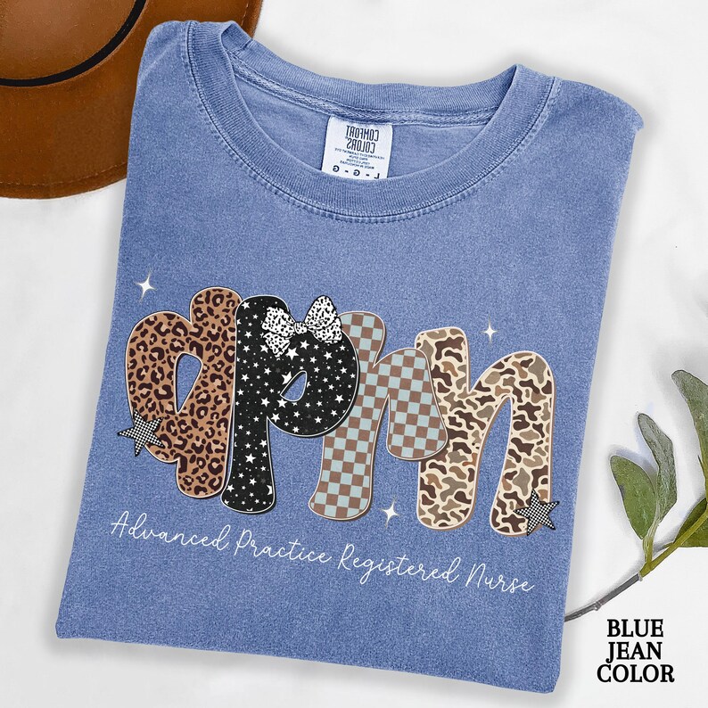 May include: A blue jean coloured t-shirt featuring the letters "APRN" in a decorative style. The letters are patterned with leopard print, stars, and a checkered design. The text "Advanced Practice Registered Nurse" is below the letters.