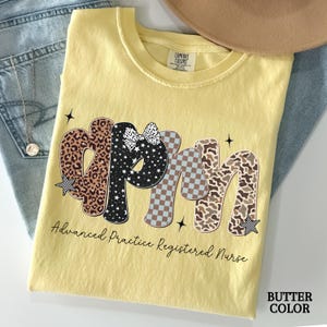 May include: A yellow t-shirt with the text "APRN" in various patterns, including leopard print, black and white stars, and checkered patterns. The words "Advanced Practice Registered Nurse" are printed below. The shirt is folded on top of blue jeans.