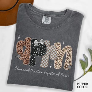 May include: Grey t-shirt featuring the letters "APRN" in a decorative style. The letters are patterned with leopard print, stars, and a checkered design. The text "Advanced Practice Registered Nurse" is printed below the letters.