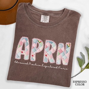 May include: Espresso-colored t-shirt with the letters APRN in a patchwork design. The letters are outlined with a scalloped edge and feature a mix of pink, blue, and green floral and checkered patterns. The text "Advanced Practice Registered Nurse" is below.