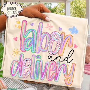 May include: An ivory t-shirt featuring the text "labor and delivery" in large, colorful, swirled letters. The design includes small graphic elements such as yellow stars, a sun, pink hearts, a butterfly, and a blue cloud scattered around the text.