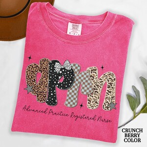 May include: A cerise t-shirt featuring the letters "APRN" in diverse patterns, including leopard print, stars, and checkered designs. The text "Advanced Practice Registered Nurse" is below. The shirt is a "Crunch Berry Color".