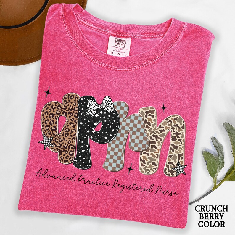May include: A cerise t-shirt featuring the letters "APRN" in diverse patterns, including leopard print, stars, and checkered designs. The text "Advanced Practice Registered Nurse" is below. The shirt is a "Crunch Berry Color".