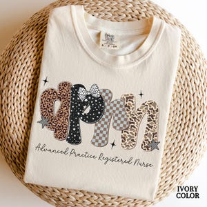 May include: Ivory-colored t-shirt with the letters "APRN" in a decorative font. The letters feature leopard print, stars, a bow, and a checkered pattern. The text "Advanced Practice Registered Nurse" is below the letters.