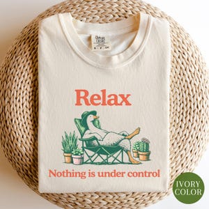 May include: Ivory-colored t-shirt with the word "Relax" in orange, featuring a cartoon duck in a cowboy hat relaxing in a chair. The text "Nothing is under control" is below the duck. The shirt is labeled "Ivory Color".
